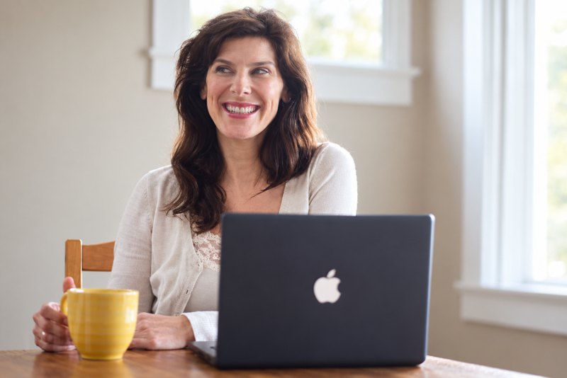 Smiling woman with a yellow mug working on a laptop at a sunny kitchen table